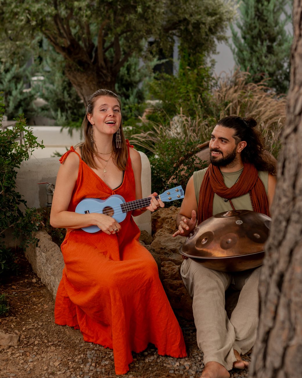 Véra singing with a ukulele, Emre playing handpan beside her on a stone bench