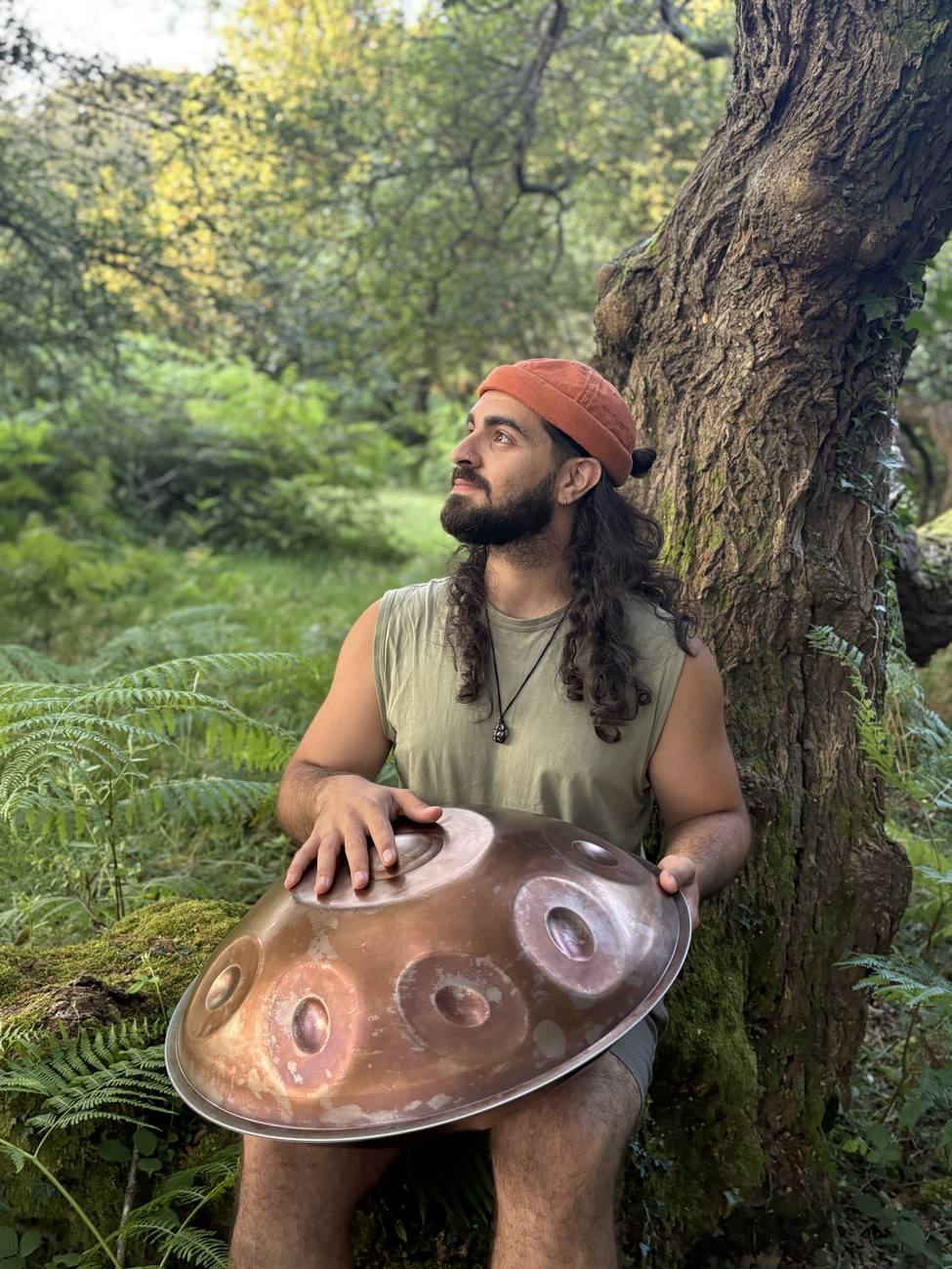 Emre with a copper handpan, leaning against a tree, looking up